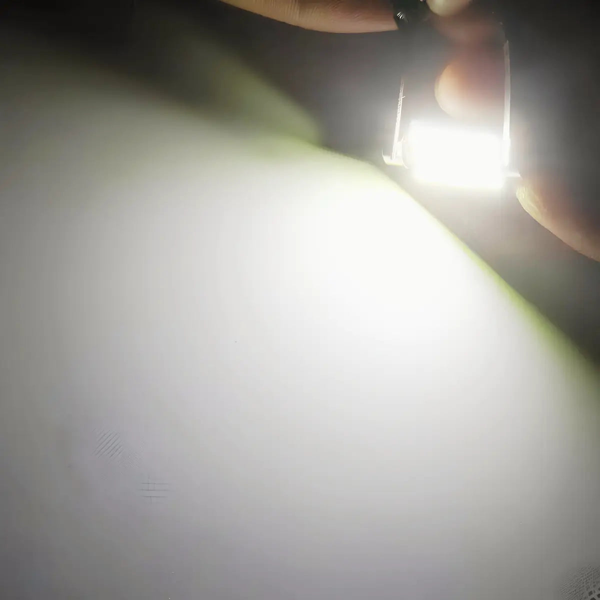 A close-up shot of a hand holding a small rectangular light source, casting a bright white beam of light onto a plain white surface.